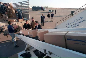 Workers handle cardboard boxes beside an aircraft stairway marked Gaza International Airport on the apron.