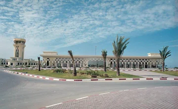 Wide view of the intact airport terminal and control tower behind landscaped grounds and palm trees.